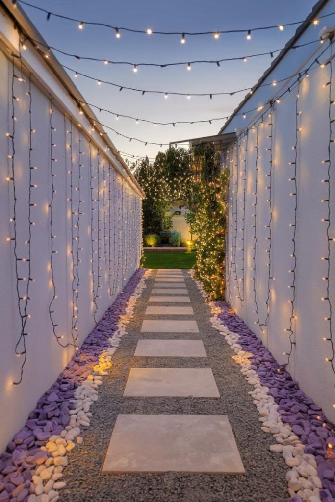 A narrow path with stepping stones is flanked by walls decorated with fairy lights. Purple and white stones line the floor. The walkway leads to a garden with trees and bushes.