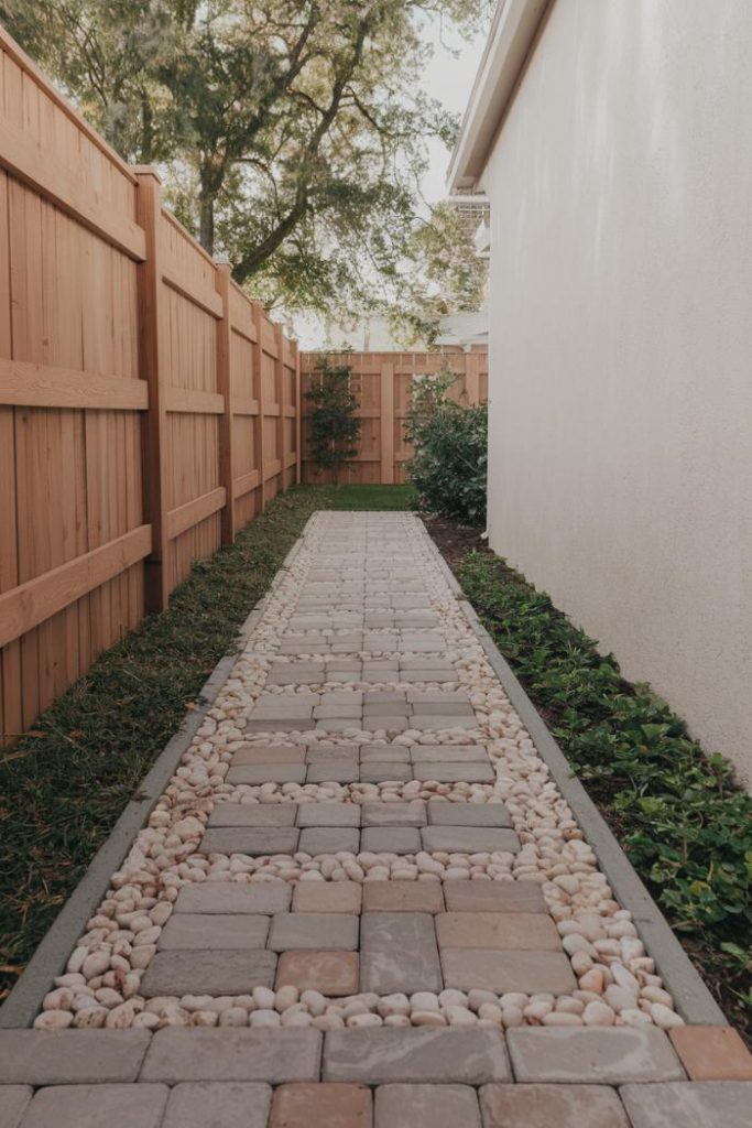 A narrow garden path features a grid of square stone tiles with moss in between. Flanked by beige walls and small plants, the entrance leads to a larger stone terrace.