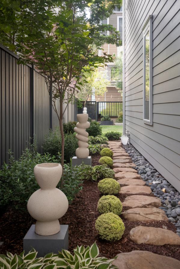 Narrow garden with stone sculptures, a row of small bushes, a stone path and a tree next to a modern house and fence.