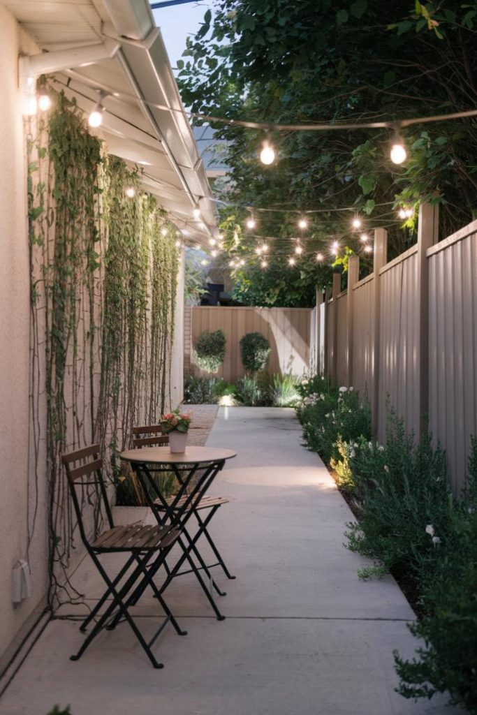 A narrow outdoor patio with fairy lights, two wooden chairs at a small table, climbing plants on a wall and bushes along a path.