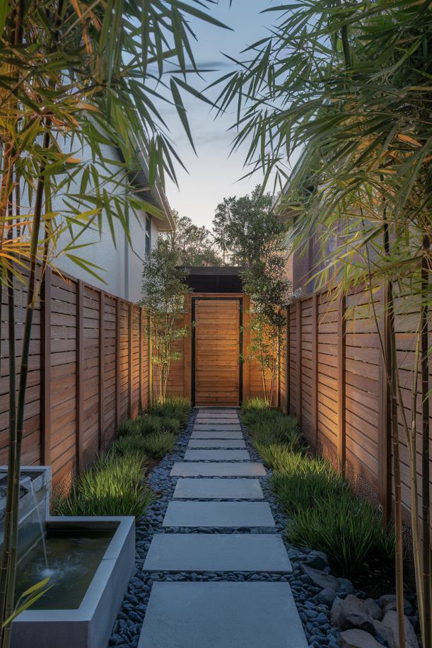 A narrow path of stone pavers leads to a wooden gate flanked by bamboo and wooden fences under a clear sky.