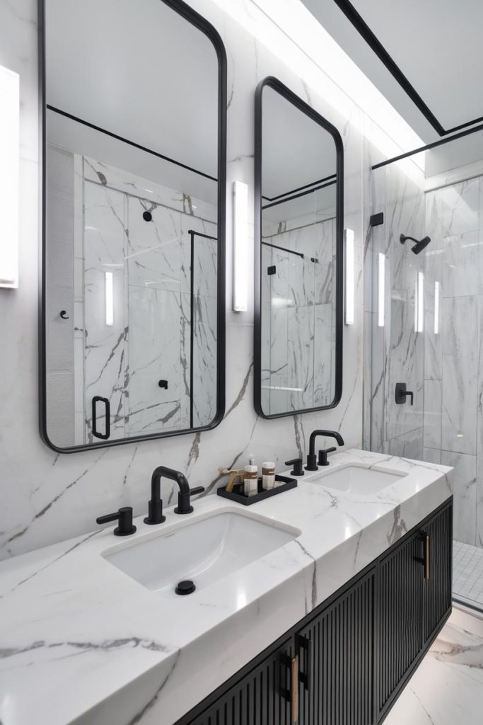 Modern bathroom with marble walls and countertops, two rectangular mirrors, two sinks with black faucets and a walk-in glass shower.