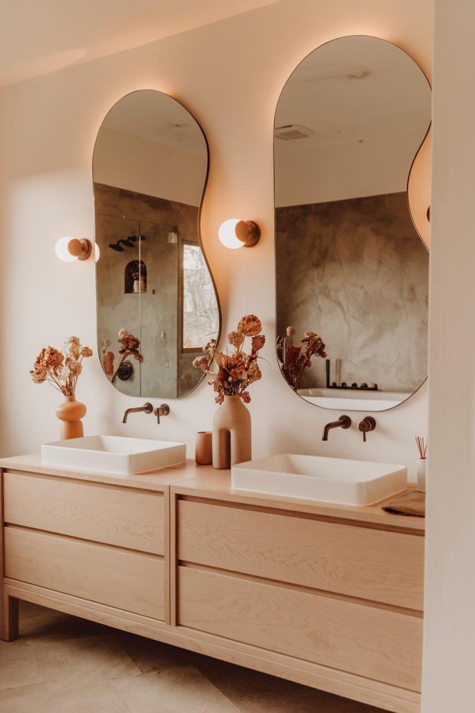 A modern bathroom with two sinks, oval mirrors and decorative vases. The lighting is warm and the room is decorated in natural tones.