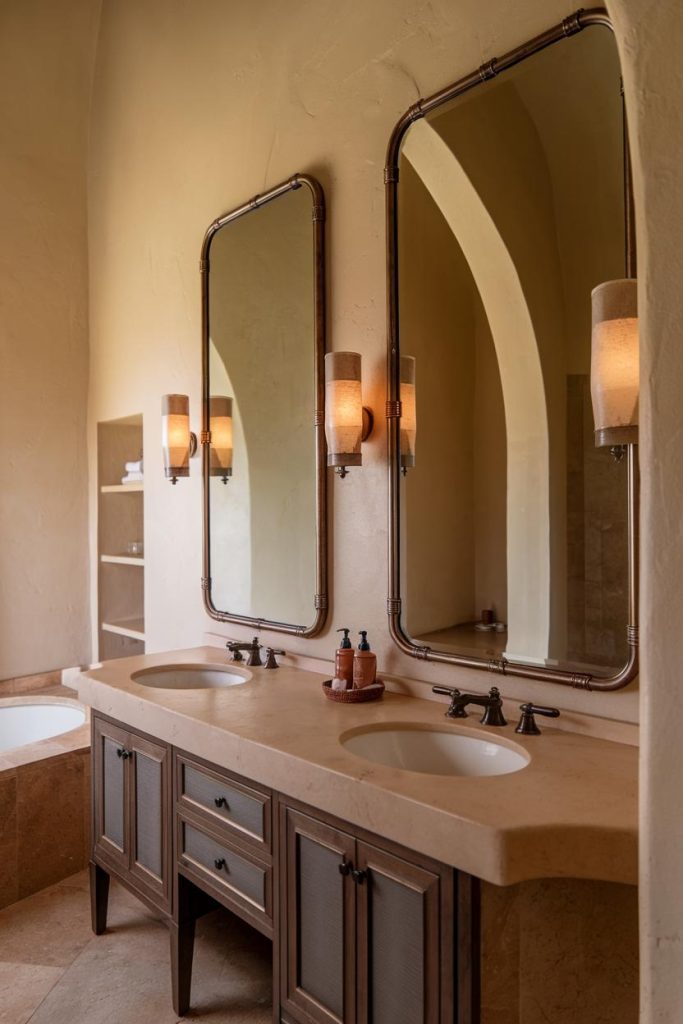 A bathroom with a double sink, two large mirrors and wall lights. A bathtub can partially be seen on the left.