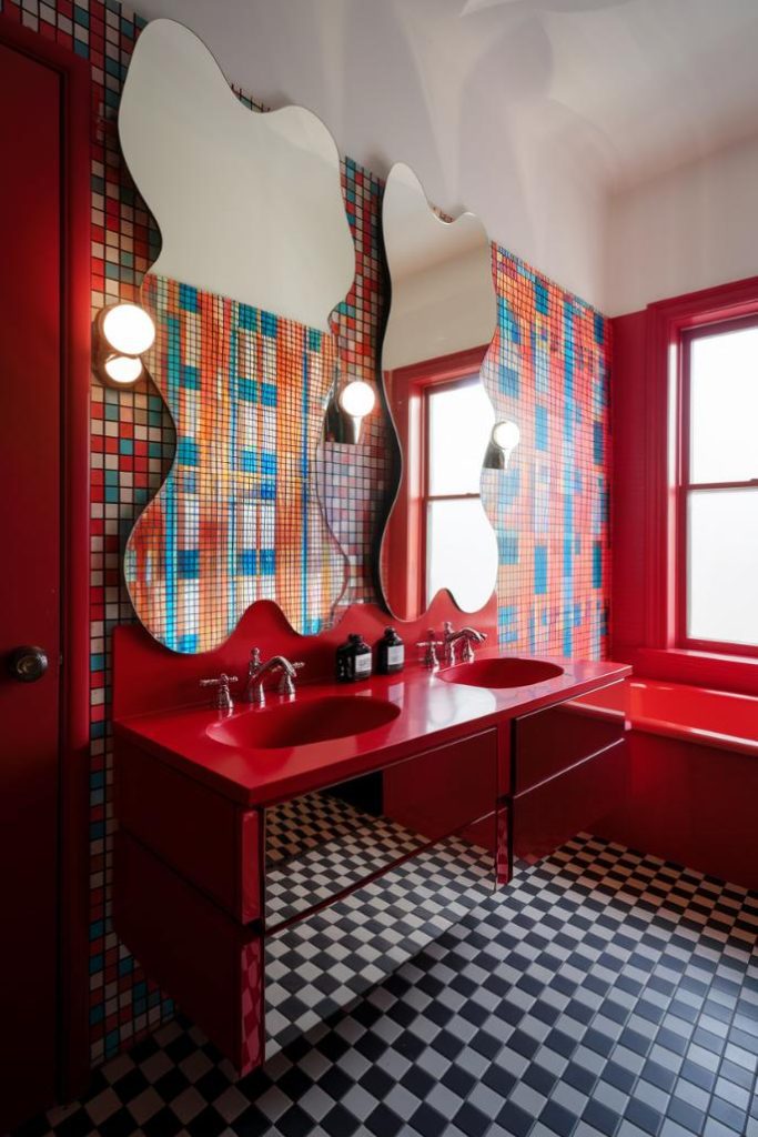 Colorful bathroom with red fittings and two wavy mirrors. Mosaic tiles on the wall and black and white checkered floor. Two round lights next to the mirrors.