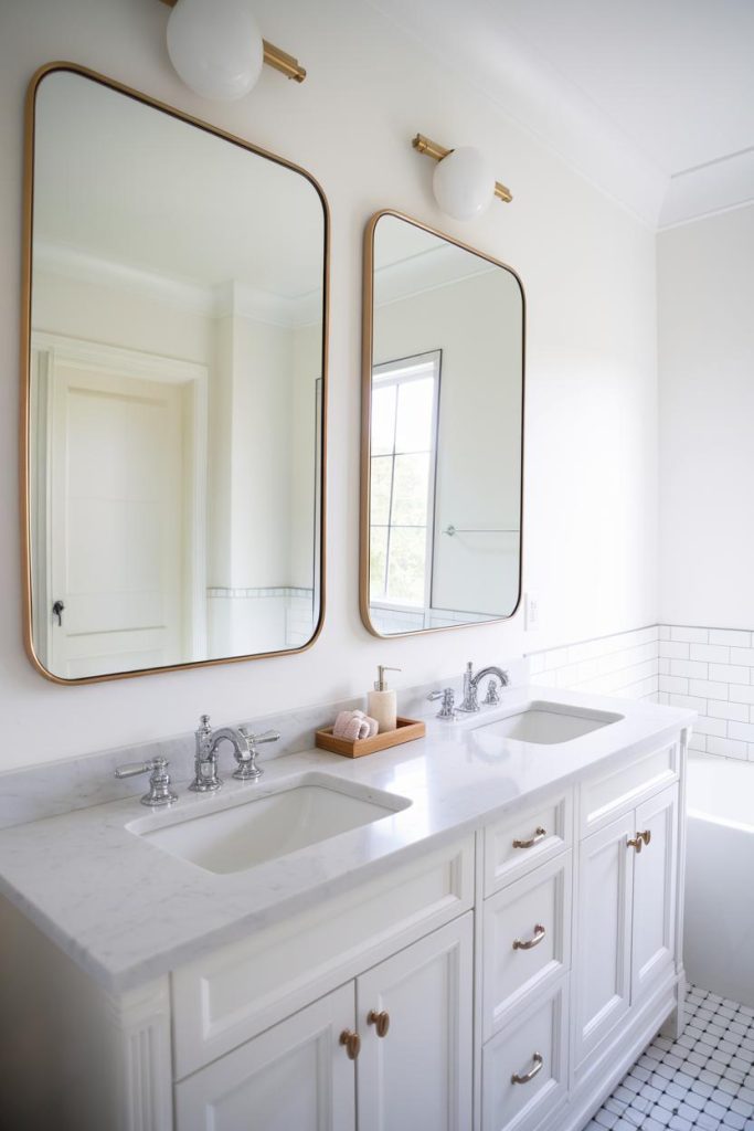 A bright bathroom features a double vanity with marble countertops, two large mirrors, and wall lights above. White cabinets have silver hardware and the floor is tiled.