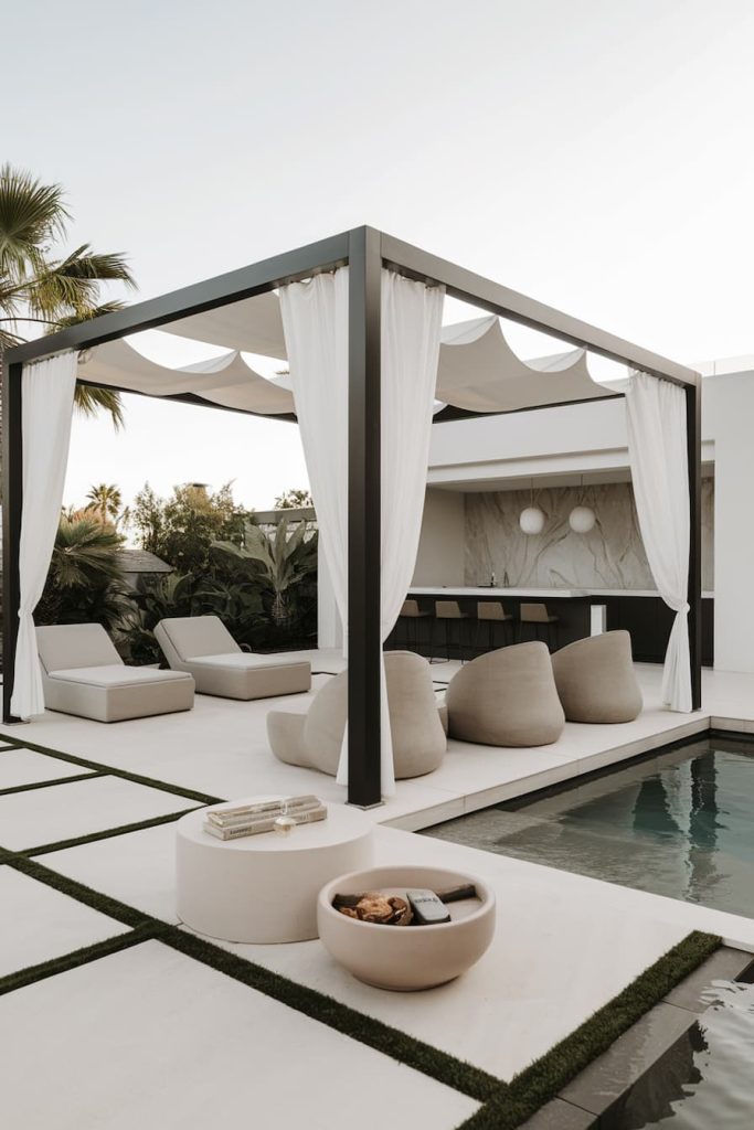 Outdoor patio with a canopy, white curtains, lounge chairs and a modern fire pit next to a pool. Minimalist design with lots of greenery in the background.