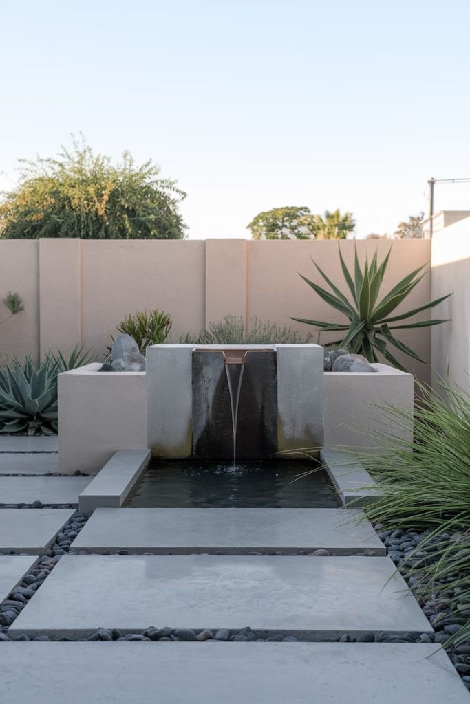 Modern garden with a rectangular fountain surrounded by large square stepping stones and desert plants in front of a high beige wall.