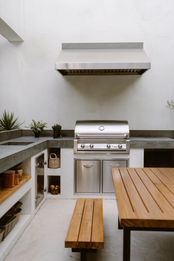 Outdoor kitchen with stainless steel grill, wooden table with bench and concrete countertops surrounded by small potted plants.