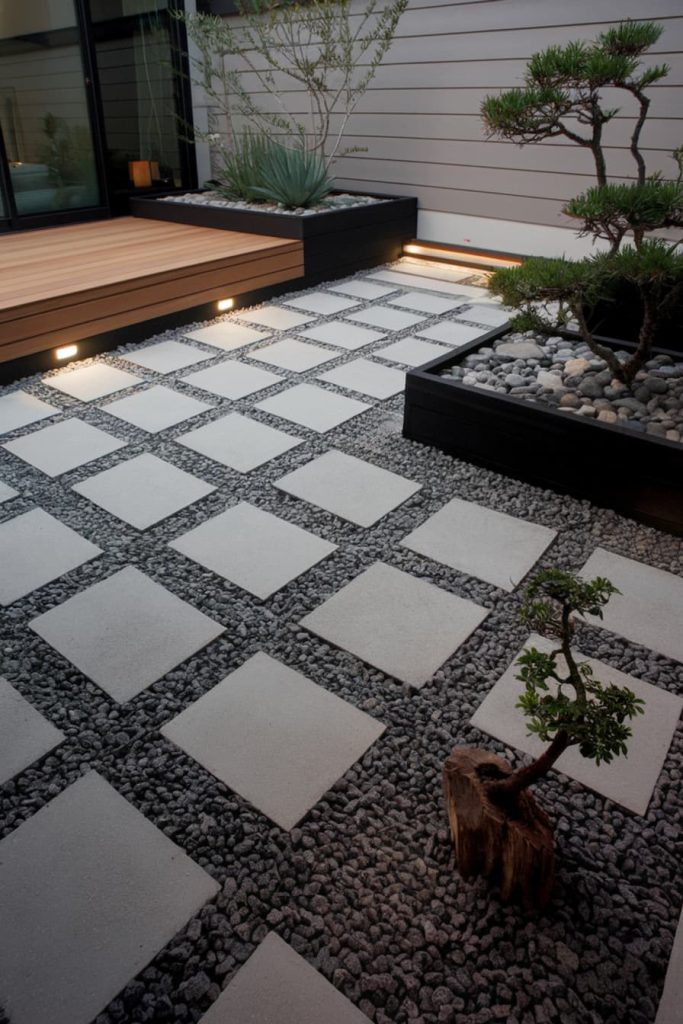 A modern patio with square stone tiles, gravel in between, a small tree in a planter, a bonsai tree and wooden patio steps leading to a sliding glass door.