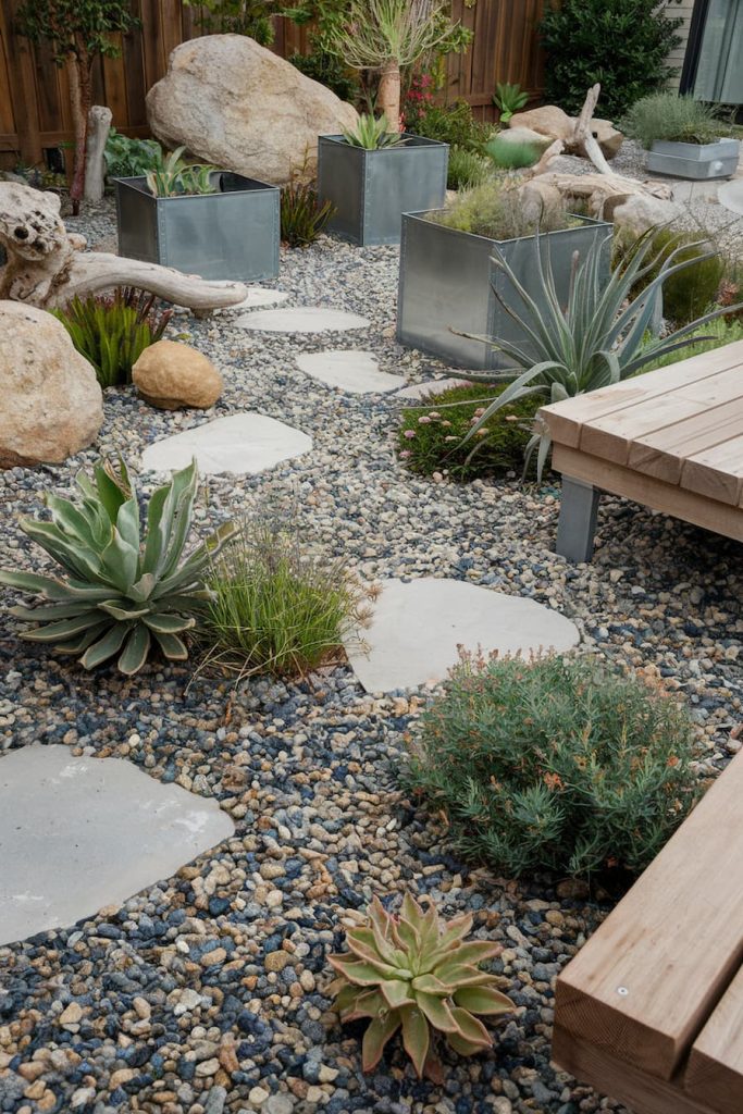 A garden of succulents in metal planters surrounded by stones and pavers, with wooden benches to the right.