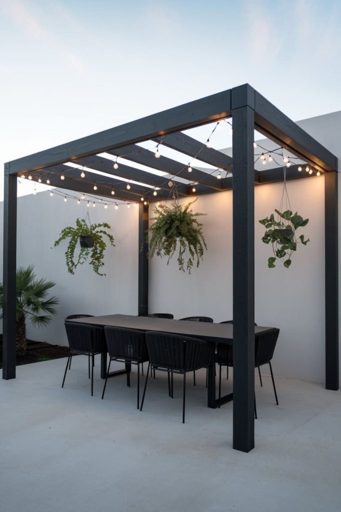 Outdoor dining area with black pergola, hanging plants, fairy lights and long table with black chairs on concrete patio.