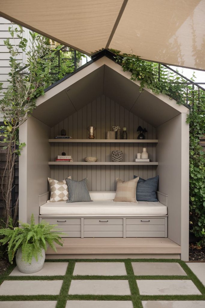 Cozy outdoor corner with built-in bench, cushions and shelves with decorative items. Surrounded by greenery and a large parasol.