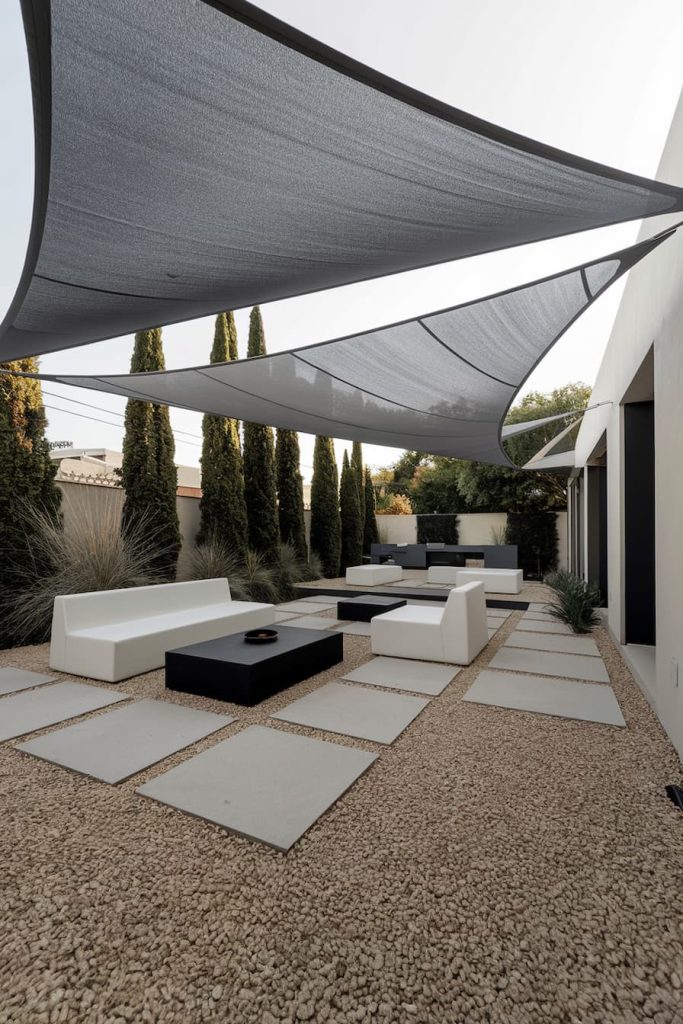 Modern outdoor patio with geometric design, light gray shade sails, white and black furniture, stone path and tall trees in the background.