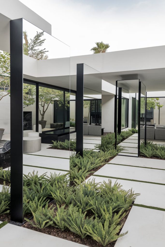 Modern outdoor patio with clean lines, mirrors, green spaces along the concrete paths and seating areas with large glass windows.