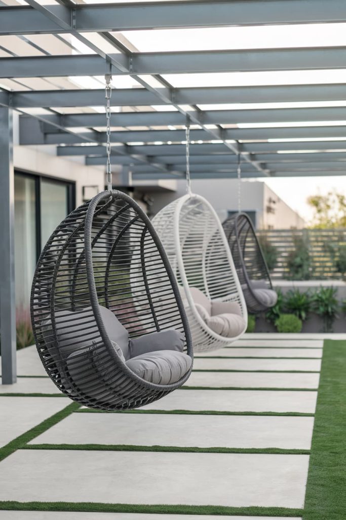 Three hanging egg chairs with cushions on a modern terrace with a pergola and beautifully landscaped surroundings.