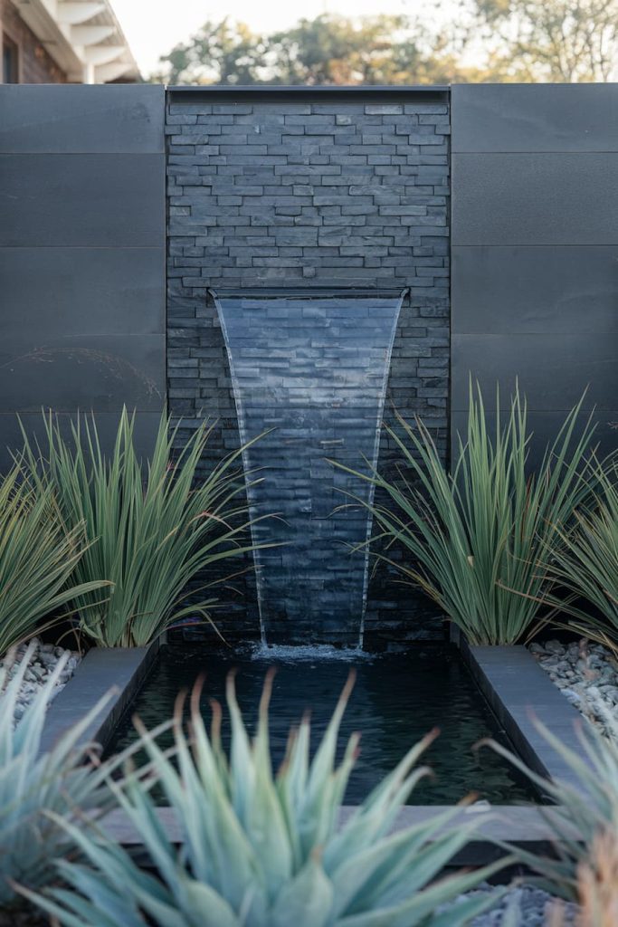 A modern water feature with a slate brick backdrop flanked by ornamental grasses and plants, with water flowing into a rectangular pond.