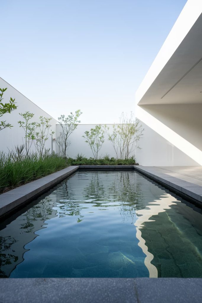 A rectangular pool with clear water is surrounded by minimalist white walls and greenery under a clear blue sky.