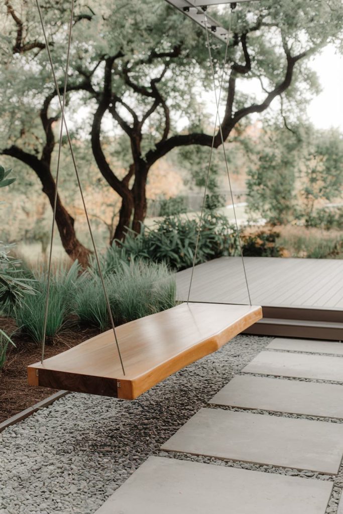 A wooden swing hangs in a garden surrounded by trees and greenery, with stone steps leading to a patio.