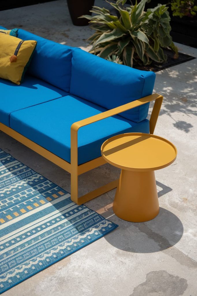 Outdoor seating area with a blue upholstered sofa, a yellow side table, a yellow cushion and a blue patterned rug on a concrete surface, near green plants.