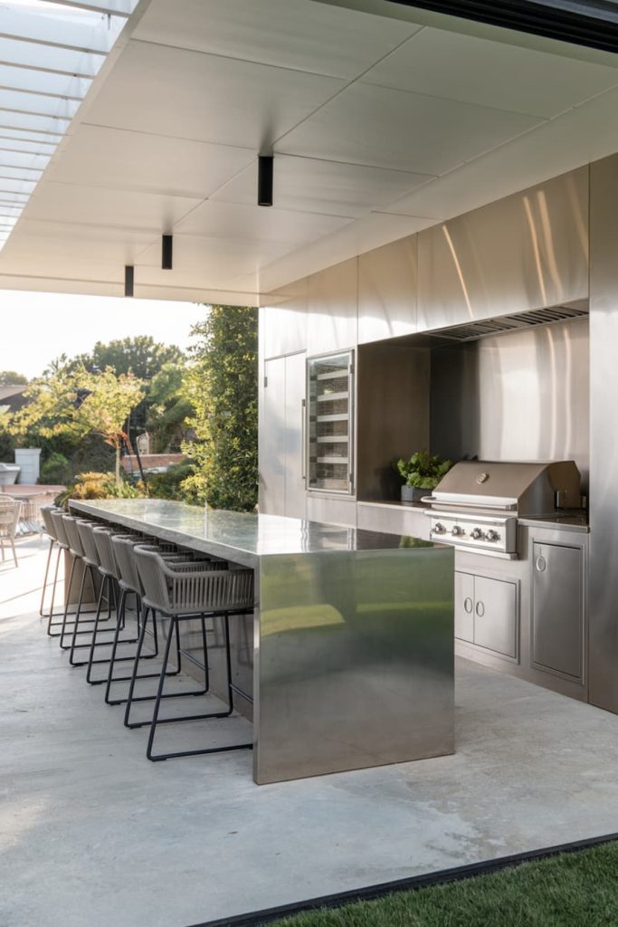 Modern outdoor kitchen with elegant stainless steel grill and matching cabinets, an island with bar stools and garden views.