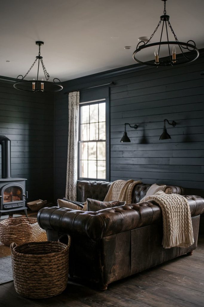 Cozy living room with dark wood paneling, brown leather sofa, knitted blankets, wood stove and two lamps. Large window and basket add to the rustic decor.