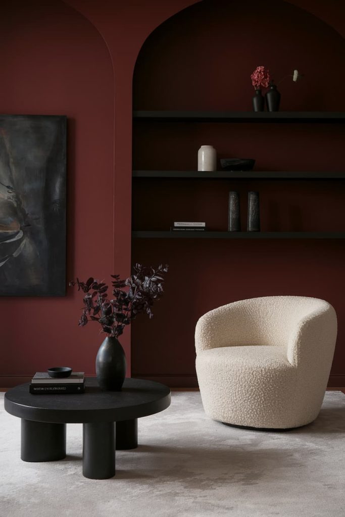 A cozy beige armchair in a dark red room with a black shelf and a round black table on which there is a vase with dark leaves and a book.