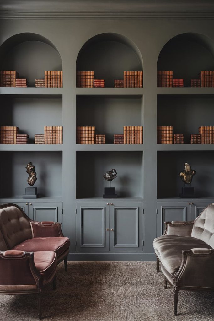 A room with gray bookshelves with books and sculptures. Two armchairs, one pink and one gray, stand in front of the shelves on a carpeted floor.