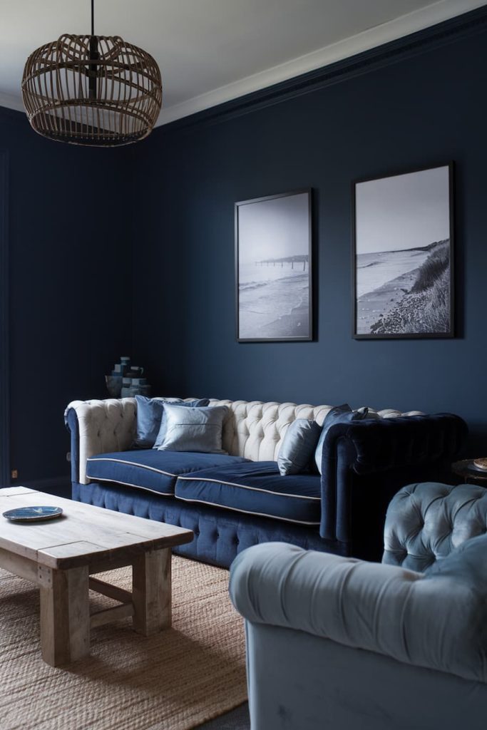 A modern living room with dark blue walls, a blue and white tufted sofa, two black and white framed pictures, a wooden coffee table and a wicker lamp.