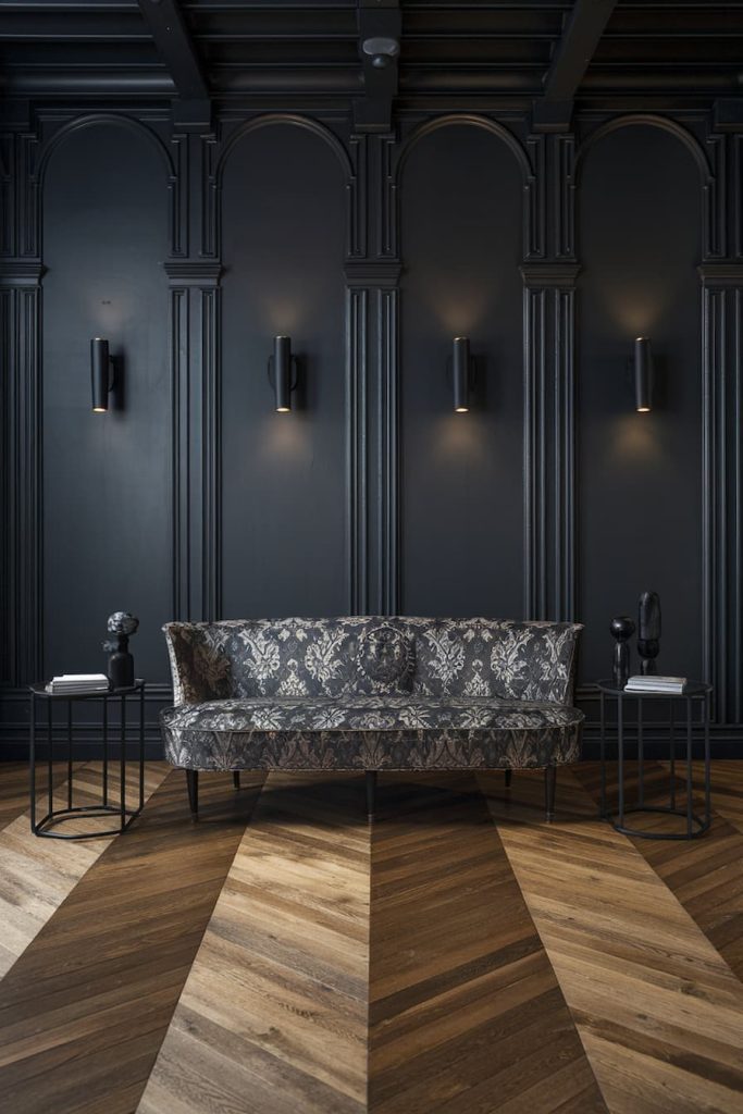 A patterned sofa in front of a dark paneled wall with five sconces. Two side tables with decorative objects stand on a geometric wooden floor.