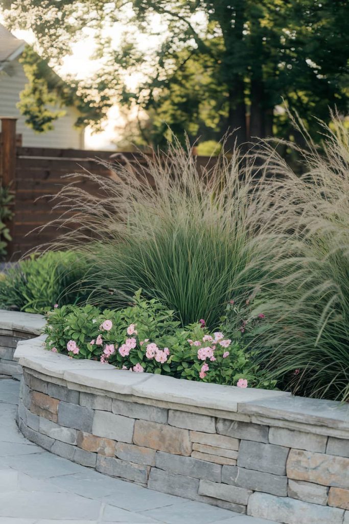 Rock garden wall with pink flowers and ornamental grasses in front and trees in the background.