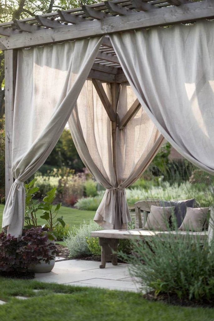 A wooden pergola with sheer curtains surrounded by lush greenery and a bench underneath on a paved area.