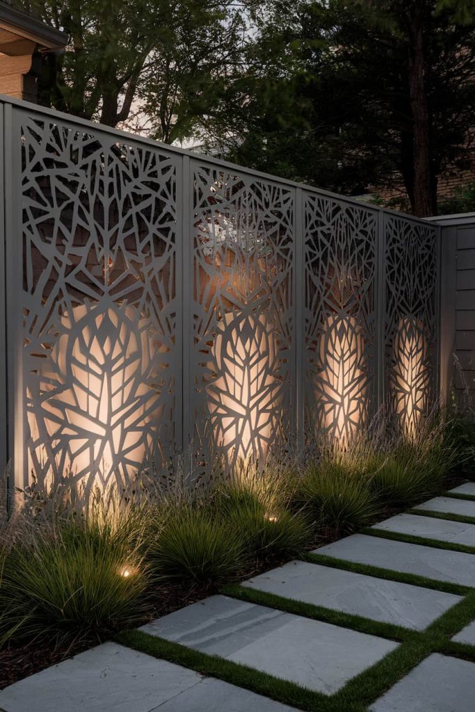 Metal panels with geometric cutouts are lit from behind, creating intricate light patterns. They line a landscaped path with rectangular stone tiles and grass. Trees and a wooden fence can be seen in the background.