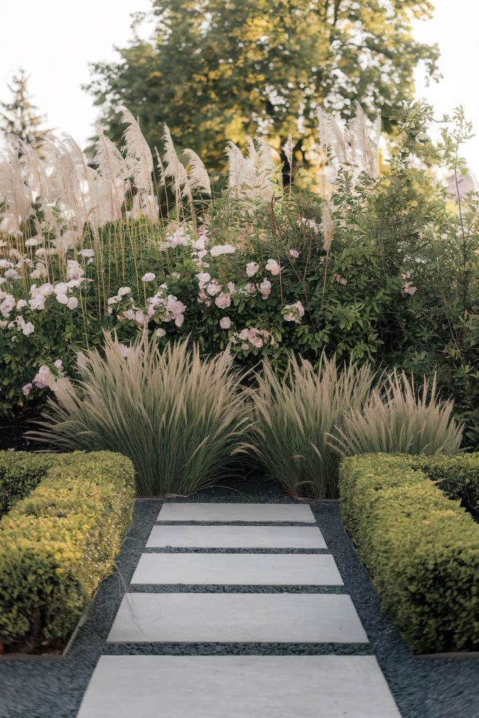 A manicured garden with a stone path flanked by trimmed hedges, ornamental grasses and blooming flowers leading into lush greenery.