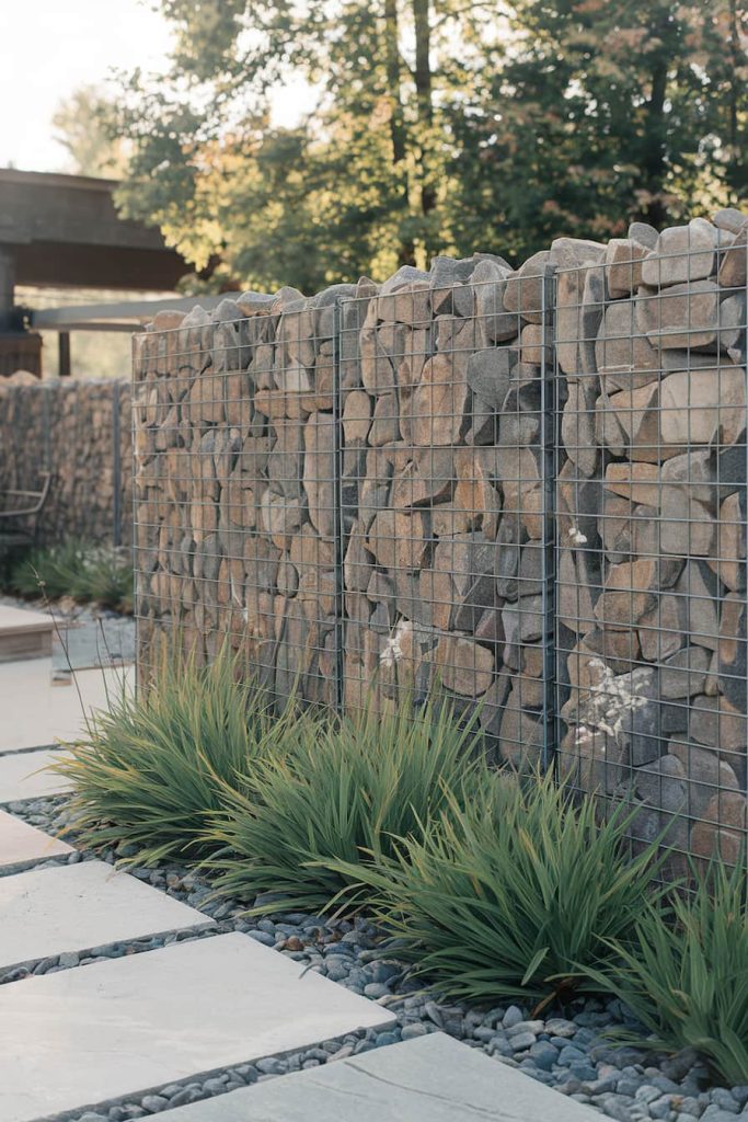 Gabion wall filled with stones along a path with rectangular stone slabs and green plants. Trees and a structure can be seen in the background.