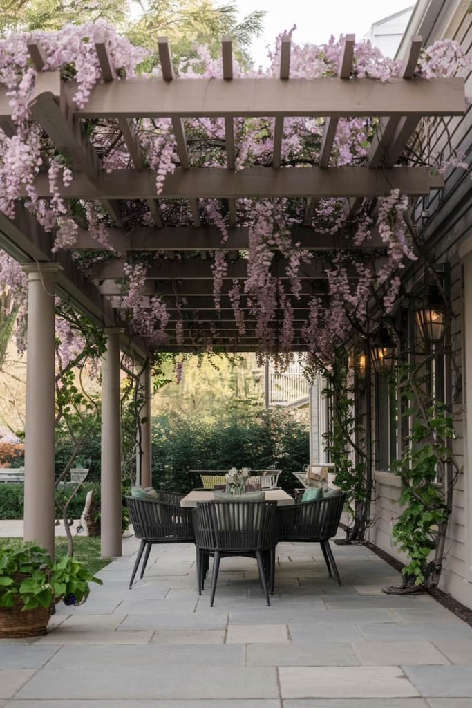 Pergola with blooming purple wisteria above a patio dining area on a stone floor.