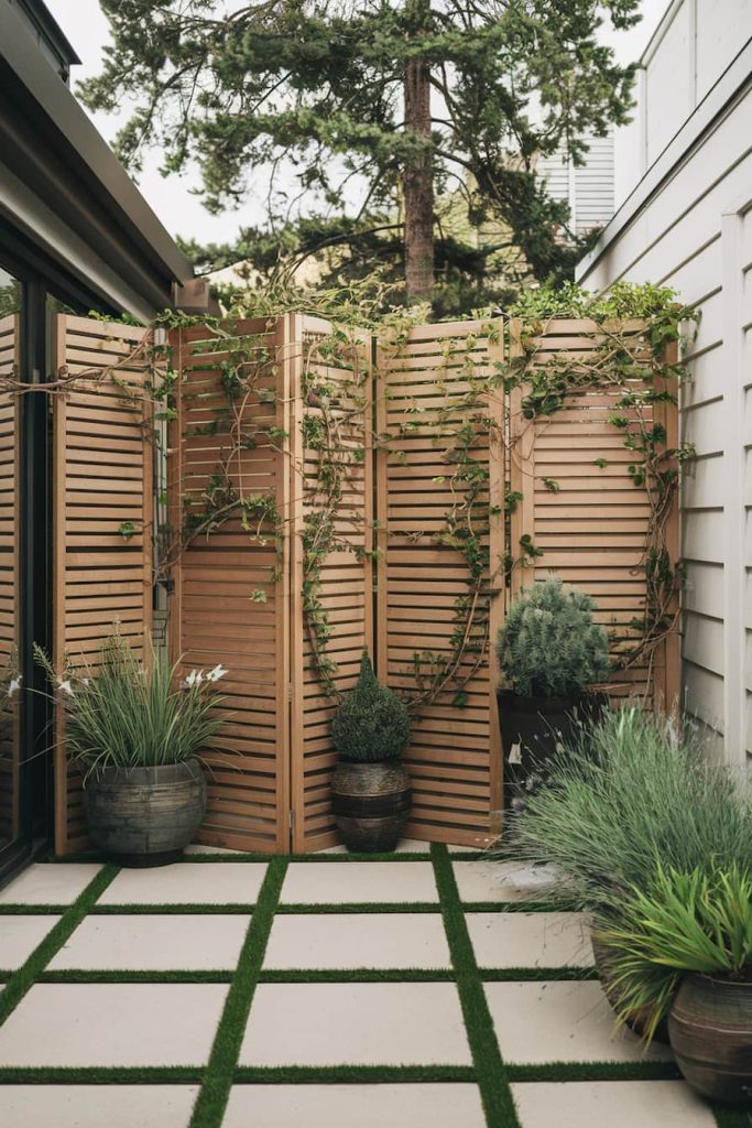 A small outdoor patio features a wooden privacy screen with climbing plants surrounded by potted plants on a tiled floor.