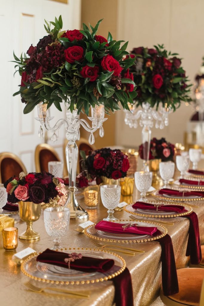 Elegant table setting with red roses, crystal glasses and gold accents on a gold tablecloth. Red napkins and candle holders add to the luxurious ambience.