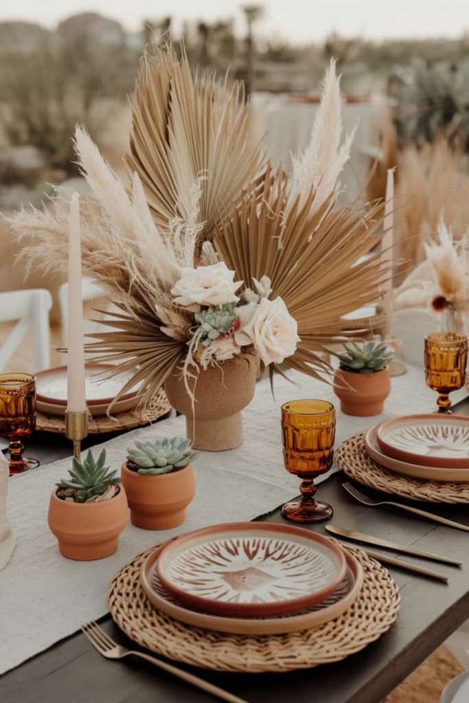 A rustic table setting featuring woven placemats, patterned plates, amber glasses, potted succulents and a central floral arrangement with dried palms and roses.