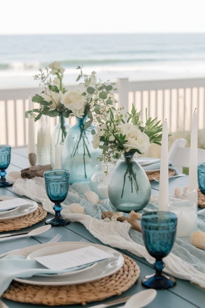 A beach table setting features blue glassware, white flowers in vases, candles and woven placemats, with a view of the sea in the background.