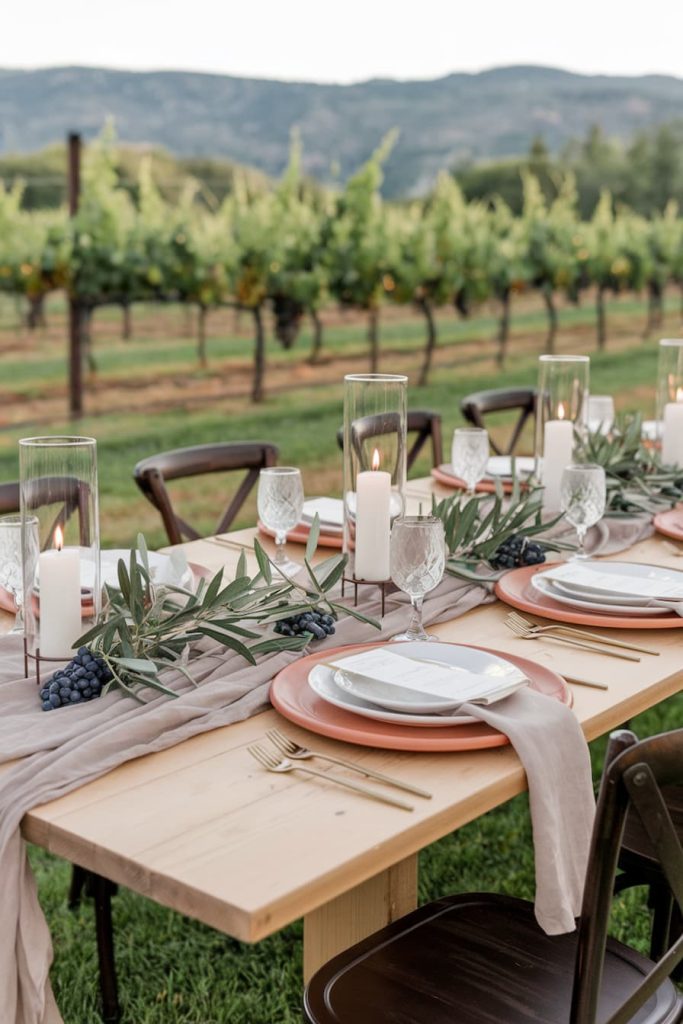 Outdoor dining table set with plates, glasses, candles and green plants, overlooking a vineyard.