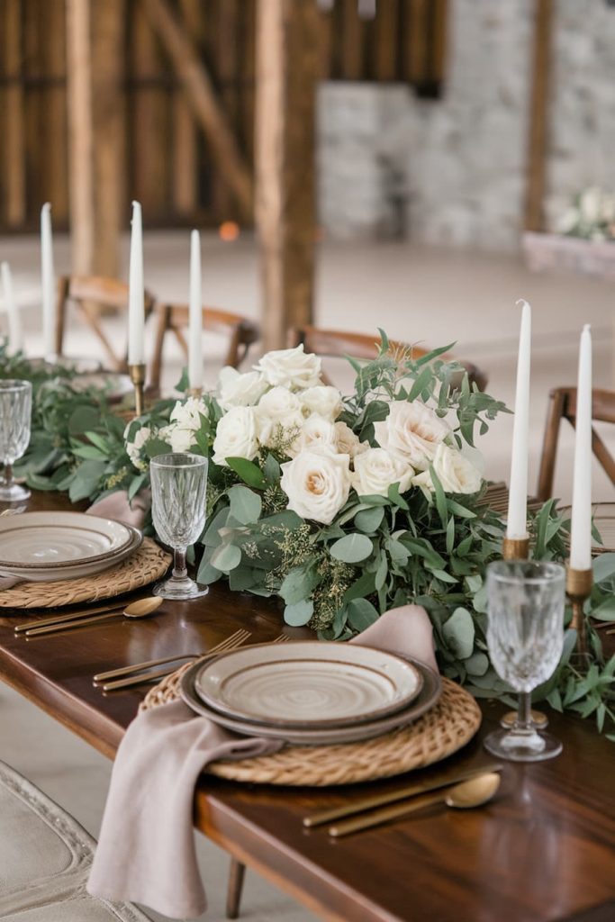 A rustic table setting with white roses and greenery, ceramic plates, glassware and white candles on a wooden table.