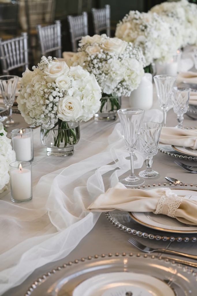 Elegant table setting with white floral arrangements, candles and crystal glasses, with cream-colored napkins on silver plates with pearl rims.