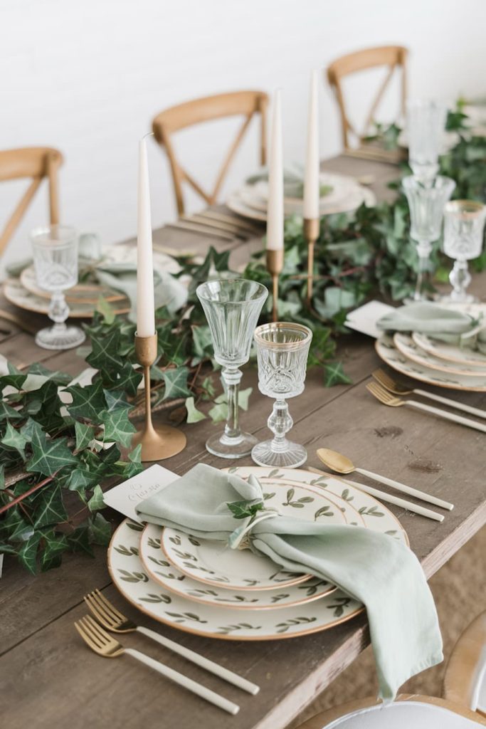 A dining table is set with green and white plates, glassware, gold cutlery and white candles. Green decorates the table.