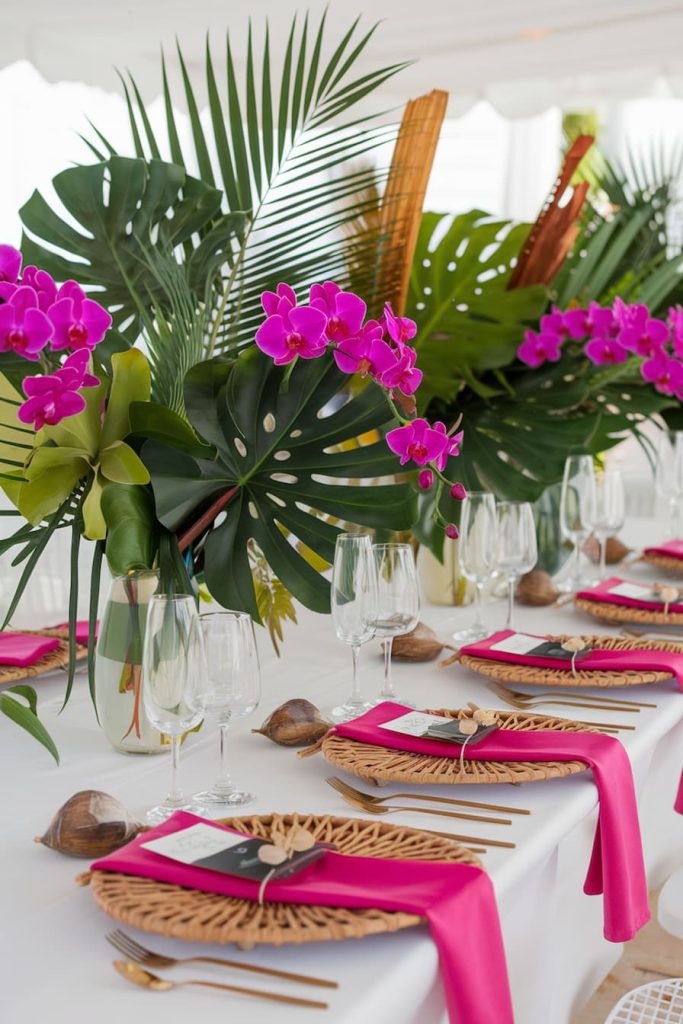 Placemat with tropical leaves, bright pink flowers, woven placemats, pink napkins and glassware on a white tablecloth.