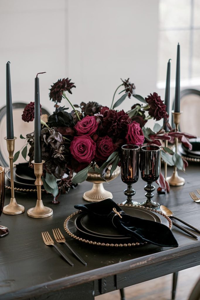 Elegant dining table with dark plates, gold cutlery, tall black candles and a centerpiece of deep red and purple flowers.