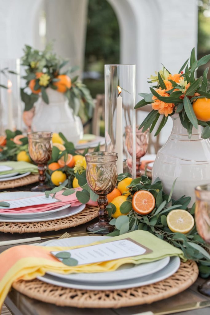 Elegant outdoor table setting with wicker tables, colorful menus, citrus fruits, amber glasses and white vases with orange flowers.