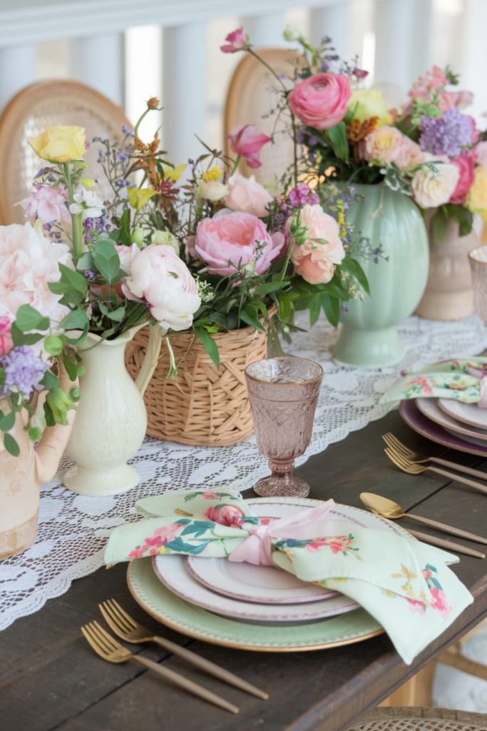 Elegant table setting with floral arrangements, pastel napkins and gold utensils on a dark wood table. Pink glassware and lace table runners complement the colorful florals.