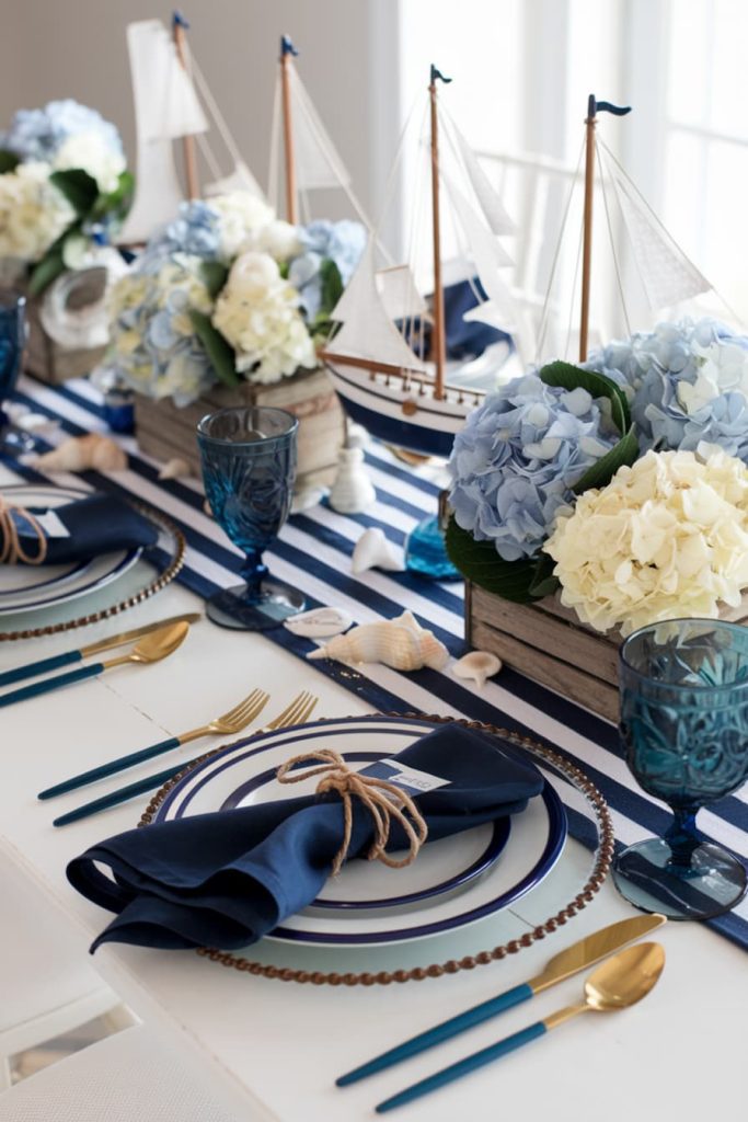 Nautical decor placemat with blue hydrangeas, model sailboats, striped runner, navy napkins and blue glassware.