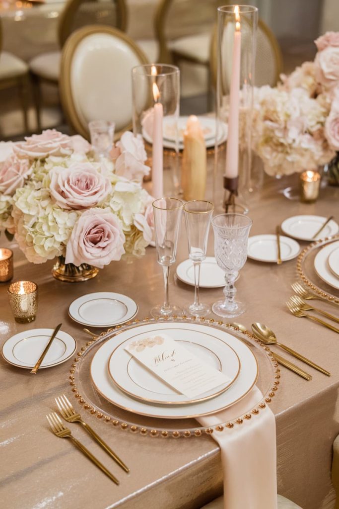 Elegant table setting with gold utensils and beaded chargers, with white and pink floral centerpieces, tall candles and glassware on a beige tablecloth.